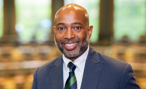 Jerlando Jackson headshot. He wears a navy blazer over a white button-down shirt. His tie is alternating stripes of navy and green. Behind him is a classroom space in Erickson Hall known as the Kiva.