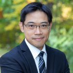 Kuo smiles toward the camera in an outside photo. Kuo wears a white button down shirt, black and gray striped tie, and a dark blazer. Kuo's black hair is cropped short. Kuo has glasses. 