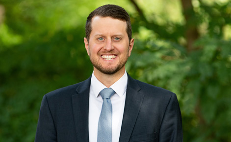 Matthew Harkey headshot. He wears a dark gray blazer over a white button-down shirt. His tie is a light blue. His brown hair is short. 