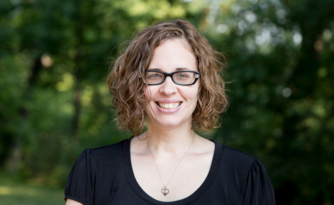 Amanda McNew. She wears a black, short-sleeved blouse. Her auburn hair is curly and cropped above her shoulders. She wears dark-rimmed glasses. 