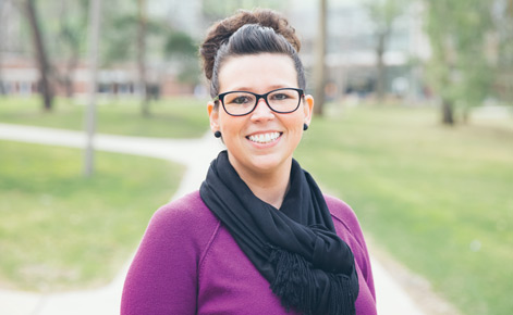 Candace Robertson. She wears a fuschia sweater, with a black scarf over top. Her brown hair is tied up in a bun. She wears dark-rimmed glasses. 