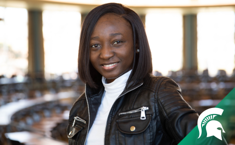 Olamide Ogungbemi wearing a black leather jacket smiling at camera. She is standing in the Erickson Hall Kiva. 