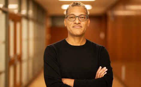 a person with curly short gray hair, wearing glasses. They are dressed in a black long-sleeve shirt and standing with their arms crossed. This person is smiling slightly and looking directly at the camera, located in a well-lit corridor that suggests an indoor office or academic environment.