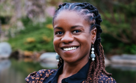 A person with long braided hair wearing a patterned blouse and large earrings, smiling in front of a blurred natural background featuring greenery and a body of water.