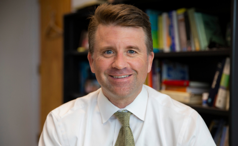 Portrait of a smiling man with short, light brown hair wearing a white dress shirt and a green patterned tie, sitting in an office with bookshelves filled with books in the background."