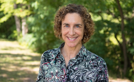 A woman with short curly hair is smiling while standing outdoors. She is wearing a patterned blouse with a mix of paisley and floral designs. The background features green trees, suggesting a natural, wooded area.