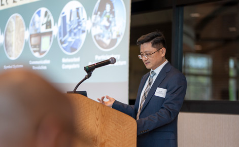 Xie stands at a podium while giving a speech; a presentation is seen, although it is blurred, behind him. Xie wears a navy suit and a brown plaid tie. 