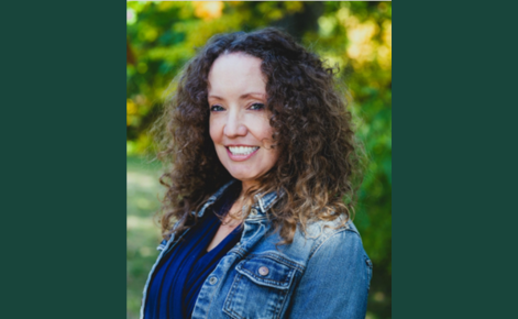 Portrait of Christina Wyman, an author. She is smiling, standing outdoors in a natural setting with green foliage in the background. She has curly hair and is wearing a denim jacket over a dark-colored top.