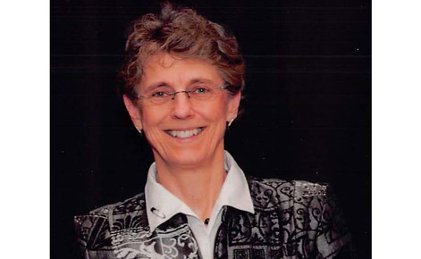 Feltz smiles toward the camera against a dark background. Feltz wears a black, gray and white blazer with a decorative pattern over a white button-down shirt. Feltz has short hair, and glasses.