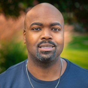 Darrius Alexander Stanley headshot. Darrius smiles toward the camera in an outside photo. Darrius wears a dark blue shirt, and a silver chain. Darrius has a goatee.