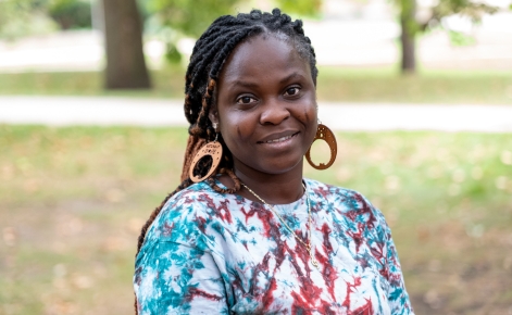 A woman standing outdoors in a park setting, wearing a multicolored tie-dye shirt with large circular wooden earrings. She has long braids, some of which are wrapped in gold. The background shows greenery with trees and blurred walking paths.