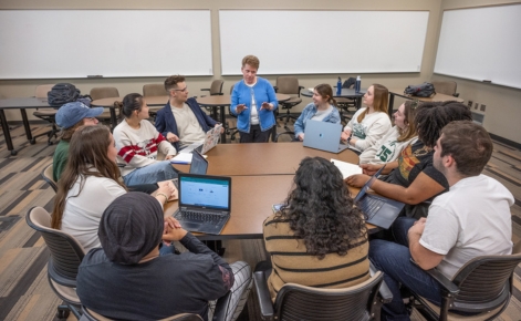 A group of diverse students is gathered around a circular table in a classroom, engaged in a discussion with a faculty member. The professor, standing at the center, is gesturing as she speaks, and all the students are focused on her. Some students are taking notes on laptops, while others have notebooks in front of them. The classroom is well-lit, and the background shows blank whiteboards and empty chairs, suggesting an interactive and collaborative learning environment.