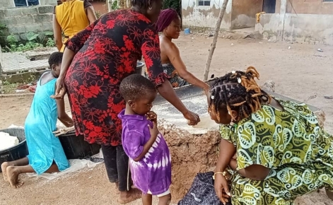 A group of women and children in an outdoor rural setting. One woman in a green patterned dress is crouching, interacting with a young boy in a purple tie-dye shirt. Another woman in a red dress is working on a task next to a large stone surface, with two other people in the background engaging in an activity, likely preparing food. The surroundings include dirt ground and a simple building.