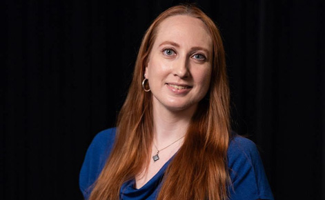 Jennifer wears a blue shirt, a necklace with a gold and blue pendant, and gold hoop earrings. Jennifer's hair is long and red and falls to the torso. Jennifer smiles toward the camera. 