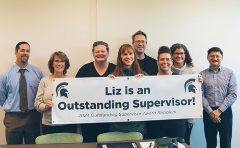 Eight Spartans gather for a group photo and hold a sign that reads: Lis is an Outstanding Supervisor! 2024 Outstanding Supervisor Award Recipient.