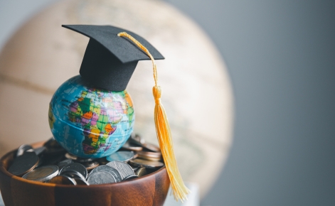 A small globe sits atop a pile of coins, symbolizing global education and investment. A black graduation cap with a yellow tassel rests on the globe, representing academic achievement and learning across the world. A blurred world map serves as the background.