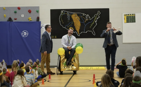 Dave Cherry sits in chair in school gymnasium while getting recognized by students and administrators.