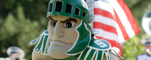 The Sparty mascot pictured with the U.S. flag blowing in the wind behind. 