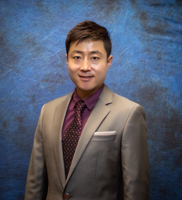 Headshot of Sanghoon Kim. He stands behind a blue and black matte background. He wears a tan suit and a dark red shirt and tie. 