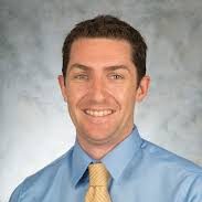 Scott Pierce headshot. Scott wears a light-blue button-down and a yellow tie. He stands against a neutral gray background. 