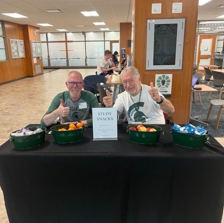 Photo of two volunteers giving a thumbs up while sitting behind a table. There are snacks for students on the table